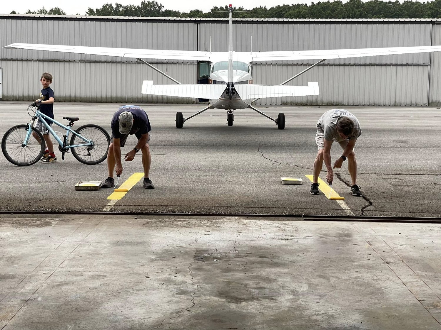 Two members painting yellow lines outside the hangar with the airplane behind them